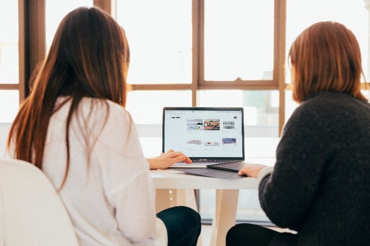 Two women sit in front of a laptop, facing away from the camera. They look like they are working together. Photo credit: KOBU Agency on Unsplash.