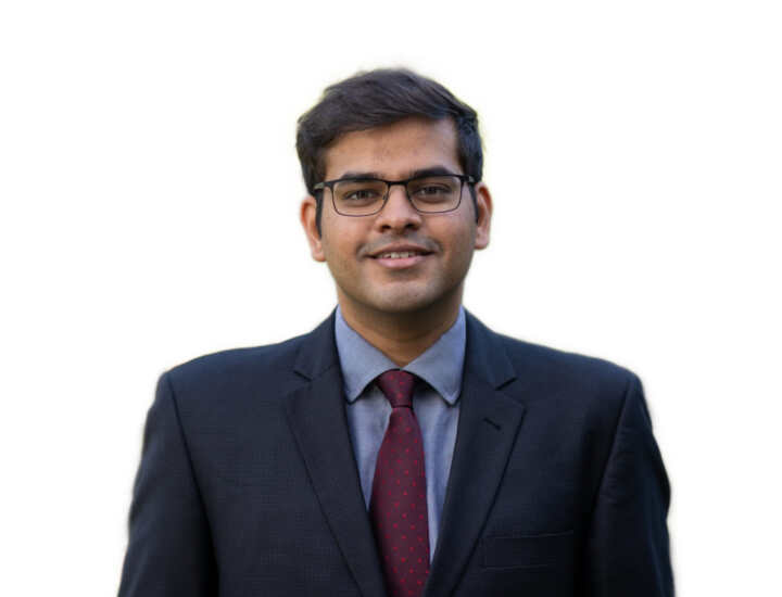 Ujjwal is smiling, he wears glasses, has short dark hair and is wearing a navy suit with a light blue shirt and a dark red tie with light red polka dots.